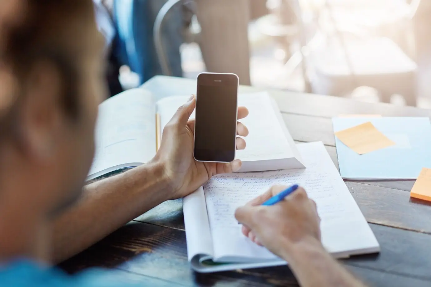 Rear view of a dark-skinned student holding a smartphone with a blank screen for your information, taking notes in a notebook while studying in a university library or canteen