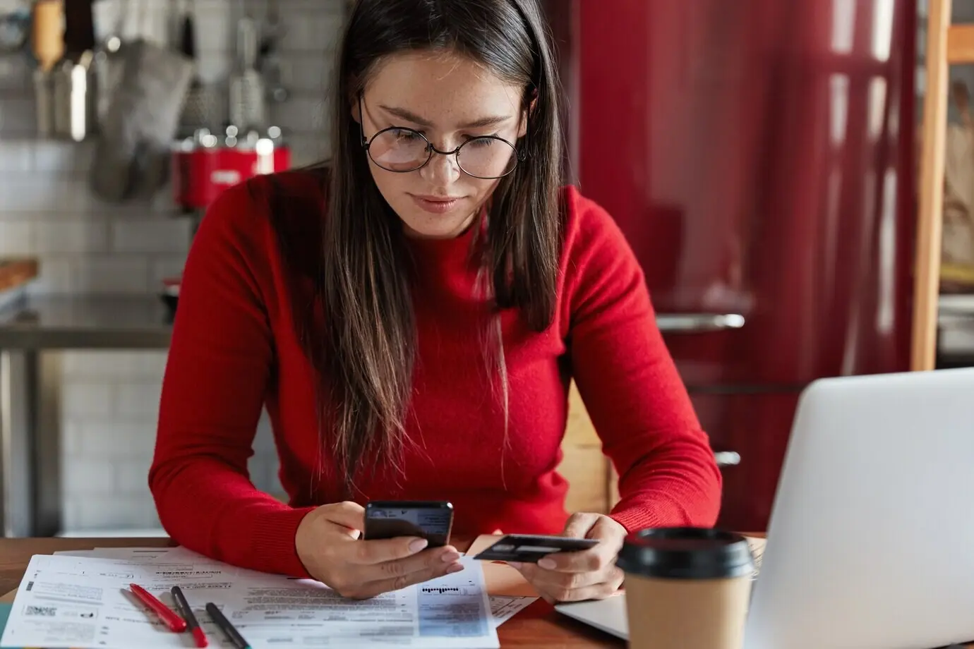 Cropped image of a freckled brunette woman in casual red clothing, holding a modern smartphone and a plastic card in her hands.